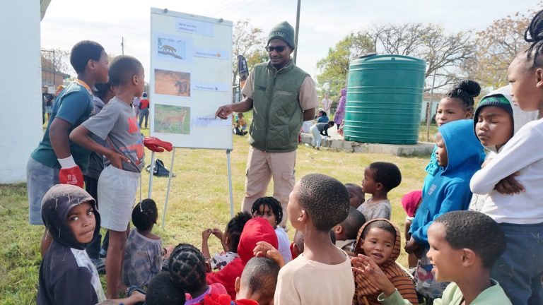 Realistic South African community members of different ages coming together outdoors at a local neighbourhood event, sharing conversation and supporting one another.